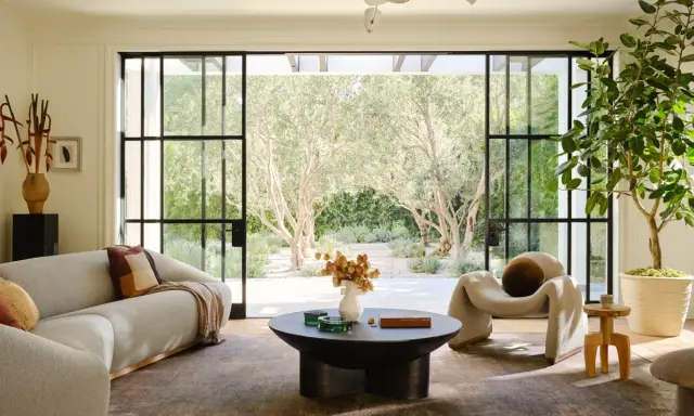 Salones modernos con sofá curvo, sillón acogedor y mesa de centro, conectados a un jardín soleado con olivos.