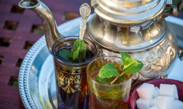 Zwei Gläser mit Minztee, garniert mit frischen Minzblättern, stehen neben einem traditionellen Teekessel und Zuckerwürfeln. Verschiedene Minze Arten verfeinern das Getränk.