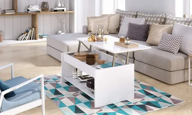 Mesa elevable pequeña blanca con compartimentos, junto a un sofá gris y una silla azul, sobre una alfombra geométrica.