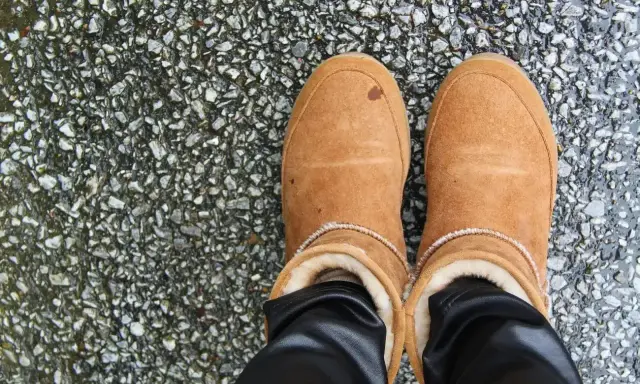 Brązowe buty z futerkiem, na których widać wytarty zamsz na butach. Stoją na mokrym, kamienistym podłożu.
