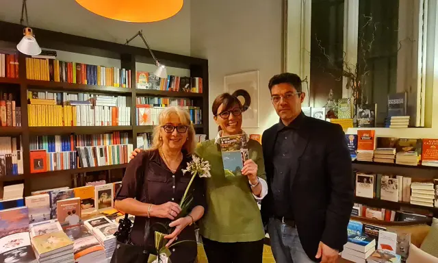 Tre persone sorridono in una libreria, una tiene in mano il libro "Cocco e Magella". Libri in ordine cronologico sugli scaffali.