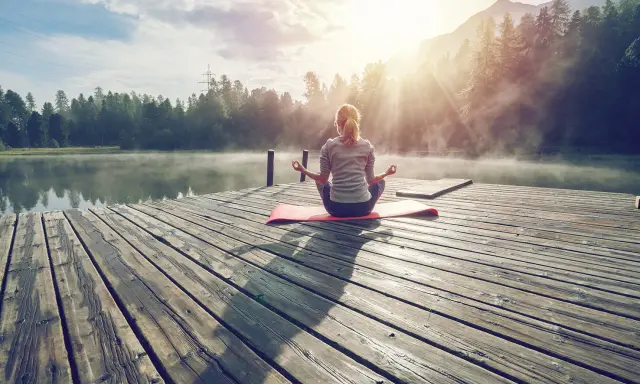 En kvinna med ryggen mot kameran sitter i meditationsställning på en brygga vid en sjö. Solen strålar genom dimman och skapar en magisk stämning. Hon verkar vara helt i nuet.