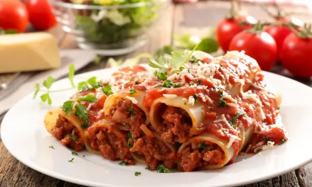 Pyszne cannelloni z mięsem mielonym, polane sosem pomidorowym i posypane parmezanem, z dodatkiem świeżej natki pietruszki.