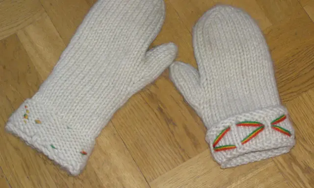 Två vita, stickade vantar med diskreta drops design-mönster i färggranna trådar på muddarna.