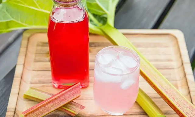 En flaska med röd rabarbersaft och ett glas med is och rosa rabarbersaft står på en träbricka. Rabarberstjälkar ligger bredvid.