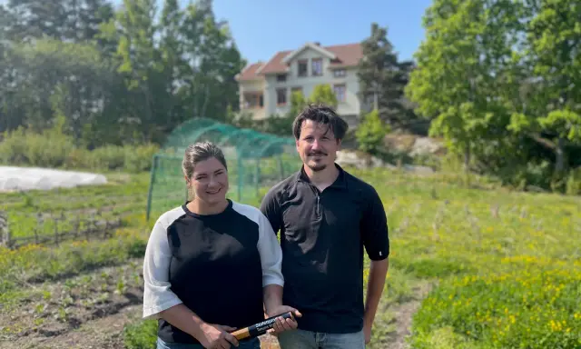 Två personer står på en åker vid Peters place Grebbestad. Bakom dem syns ett hus och grönskande träd.