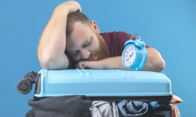 Zmęczony podróżnik śpi na walizce, budzik obok sygnalizuje jetlag.