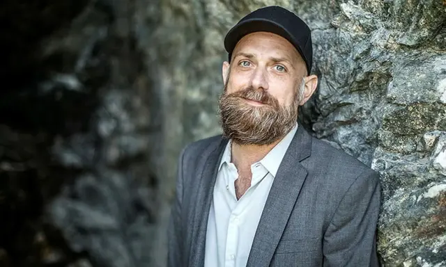 Man med skägg och keps, i grå kavaj, står mot en stenvägg. Kanske en del av en förintelsen serie på SVT.