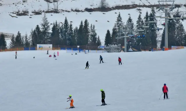 Narciarze i snowboardziści szusują po ośnieżonym stoku w Bukowinie Tatrzańskiej. W tle widać wyciąg krzesełkowy i las.