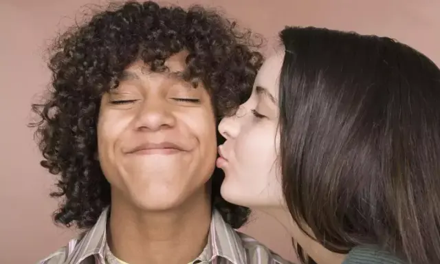 Un jeune homme aux cheveux bouclés reçoit un baiser sur la joue d'une jeune femme. Il semble ravi, les yeux fermés. Un moment de tendresse, un vrai pécho def.