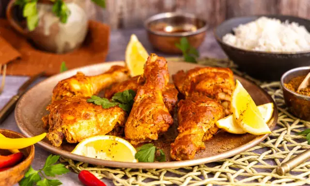Saftiges Chicken Tandoori auf einem Teller mit Zitronenspalten und Koriander. Daneben Reis und Gewürze.