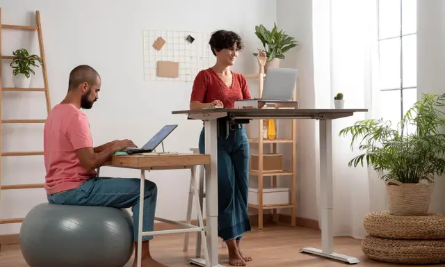 Un homme travaille sur son ordinateur, assis sur un ballon de gym, tandis qu'une femme travaille debout à un bureau réglable. Un espace de travail moderne et dynamique.
