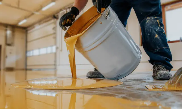 Persona con guantes vierte pintura amarilla de un cubo para pintar suelo cemento sin pulir.
