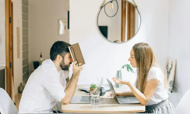 Hombre con personalidad narcisista, frustrado, se cubre la cara con un libro mientras una mujer le habla con exasperación.