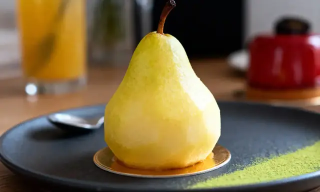 Un entremet poire, glacé et brillant, repose sur une assiette noire avec une touche de poudre verte.