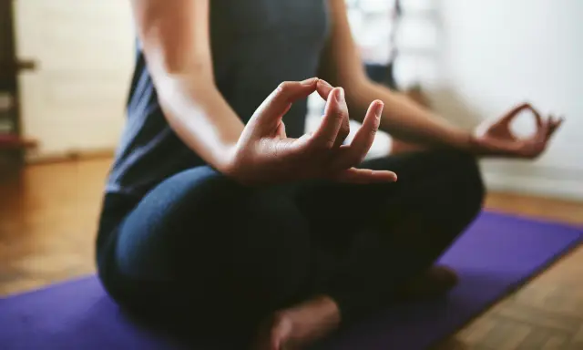 En person sitter i meditationsställning på en lila matta. Händerna vilar i knät med tumme och pekfinger sammanfogade, en vanlig gest för att visa hur man mediterar.