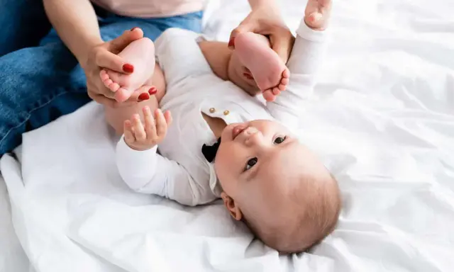 Mama delikatnie ćwiczy nóżki niemowlaka, pomagając mu przy wzmożonym napięciu mięśniowym. Dziecko leży na białym tle.