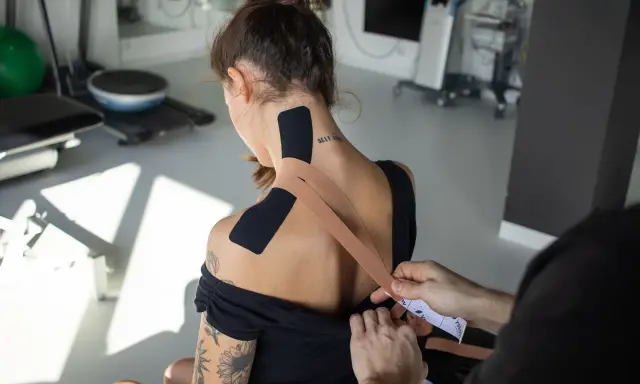 Aplikacja tejpy na szyję i barki. Dłoń nakleja taśmę kinezjologiczną na plecy kobiety.
