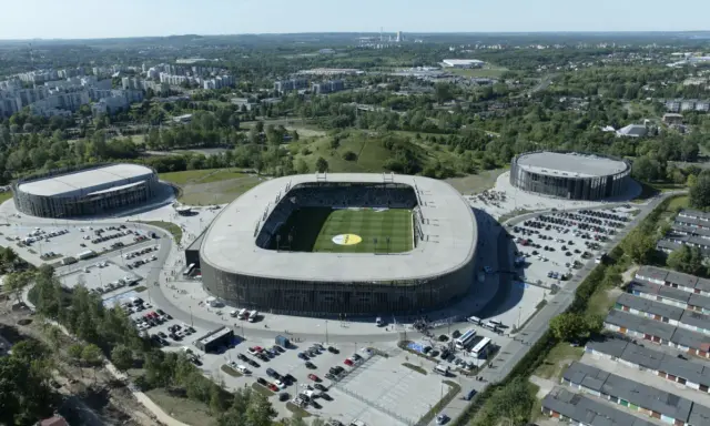 Widok z lotu ptaka na nowoczesny kompleks sportowy, w tym stadion piłkarski i dwie hale widowiskowe. To arena Sosnowiec, otoczona zielenią i parkingami.