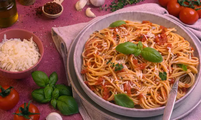 Ein Teller Spaghetti Napoli mit Tomatensauce, Basilikum und Parmesan. Daneben Zutaten wie Tomaten, Knoblauch und Olivenöl.