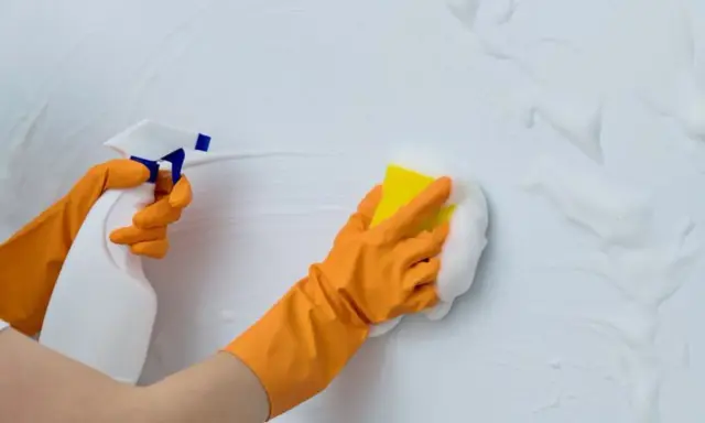 Manos con guantes naranjas limpian paredes blancas con spray y esponja. Guía práctica: como limpiar paredes blancas.