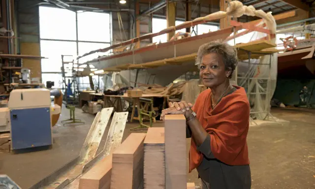 Jacqueline Tabarly, souriante, dans un atelier de construction navale, devant des blocs de bois et un voilier en cours de fabrication.