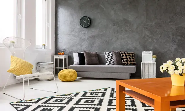 Nowoczesny salon z szarą ścianą, na której widać ciekawe efekty. Kanapa, biały fotel i geometryczny dywan tworzą przytulną przestrzeń.