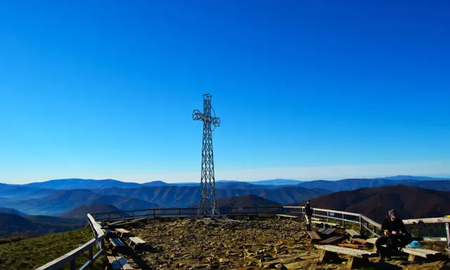 Metalowy krzyż na szczycie i turysta odpoczywający na ławce. Widok na bieszczady, gdzie niebo jest błękitne.