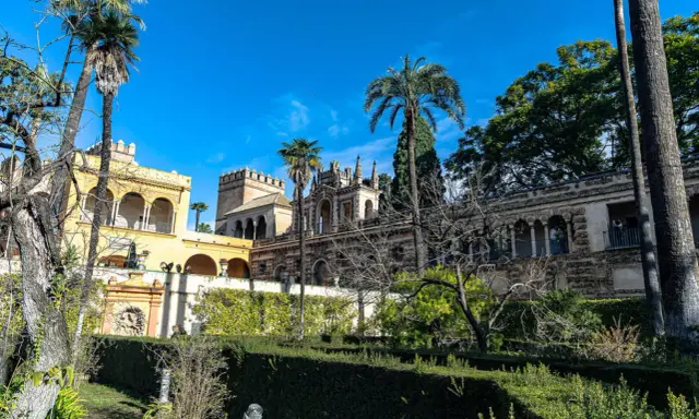 Sonniger Tag im Alcázar von Sevilla. Palmen ragen über kunstvoll verzierte Gebäude und gepflegte Gärten.