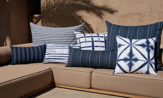 Plusieurs coussins bleus et blancs, à rayures et motifs tie-dye, sont disposés sur un canapé marron. Idée pour comment mettre un plaid sur un canapé.