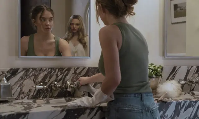 Una ragazza si pulisce il lavandino mentre un'altra la osserva riflessa nello specchio. La scena suggerisce una trama di famiglia complessa.