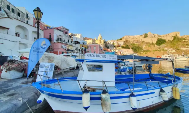 Barca bianca e blu ormeggiata, con bandiera "Pesca Turismo Isola di Procida". Case colorate sullo sfondo.