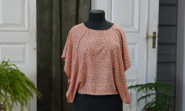 En persikofärgad blus med ett litet mönster av mörka prickar och små blommor. Blusen har en vid passform och korta, fladdermusliknande ärmar.