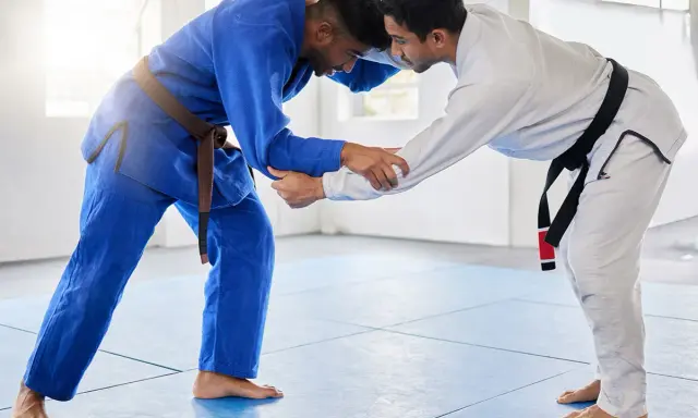 Dwóch mężczyzn w kimonach do BJJ, jeden w niebieskim z brązowym pasem, drugi w białym z czarnym pasem, ćwiczy chwyty.