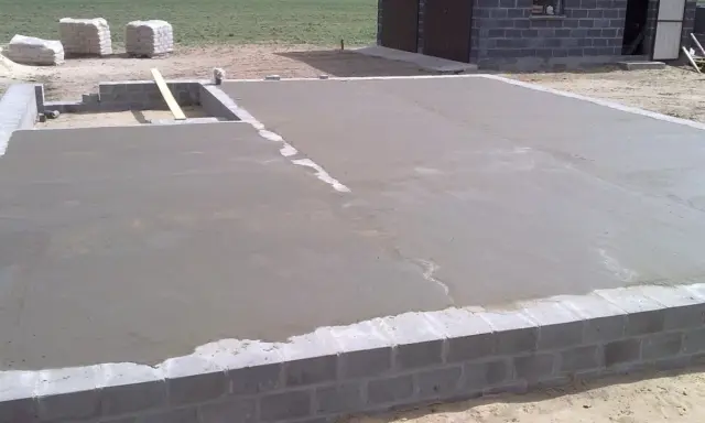 Świeżo wylany beton na chudziaka, tworzący fundament pod budowę. W tle widać materiały budowlane i fragment domu.