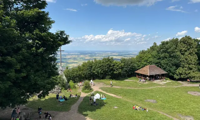 Ludzie odpoczywają na trawie i spacerują po szlakach na górze Ślęża. W oddali widać rozległą panoramę.