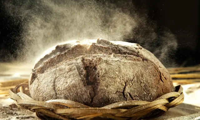 Un pain au seigle rustique, saupoudré de farine, repose dans un panier en osier. La poussière de farine danse dans la lumière, créant une atmosphère magique.