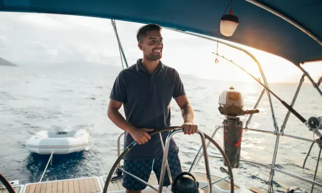 Un homme souriant est au gouvernail, prêt à barrer un bateau. Le soleil brille sur l'eau calme.