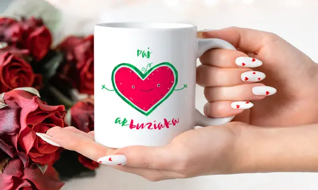 Biały kubek z zabawnym sercem-arbuzem i napisem "Daj buziaka", trzymany w dłoniach z paznokciami w serduszka, na tle czerwonych róż.