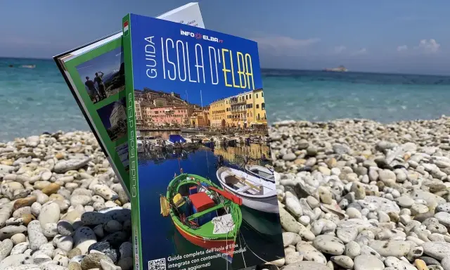 Guida turistica "Isola d'Elba" su una spiaggia di ciottoli, con il mare azzurro sullo sfondo.