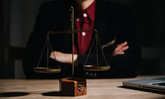 Bilancia della giustizia su un tavolo di legno, con una persona in giacca e camicia rossa sullo sfondo. Le ultime notizie sulla legge Pinto sono in arrivo.