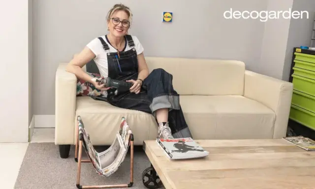Mujer con overol sentada en sofá beige, sosteniendo una herramienta. A su lado, un revistero diy hecho con cobre y tela.