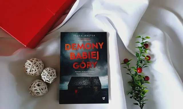 Książka "Demony Babiej Góry" Ireny Małysy, leżąca na białym jedwabiu, obok czerwonego pudełka i ozdobnych kulek. Historia, która zabiera daleko od Babiej Góry.