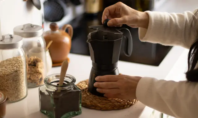 Manos preparando un café con alcohol en cafetera italiana. Hay frascos con avena y café molido.