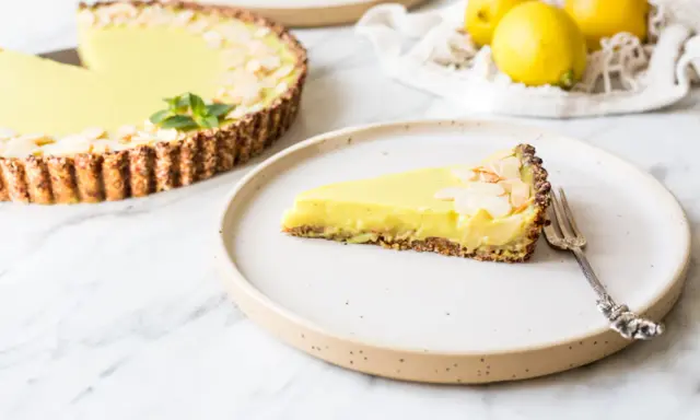 Ein Stück zitronentarte vegan mit Mandelsplittern auf einem Teller. Daneben eine ganze Tarte und frische Zitronen.
