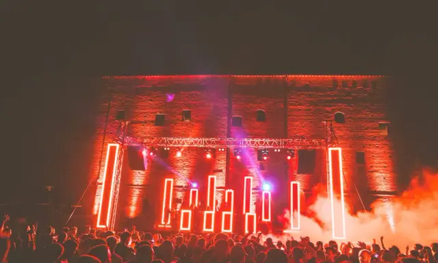 Folla in delirio al **mood festival**, illuminata da luci al neon rosse e fumo denso. La scena è un'esplosione di energia.