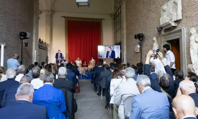 Un evento per il meeting per l'amicizia fra i popoli. Un relatore parla a un pubblico seduto, mentre un altro viene trasmesso su uno schermo.