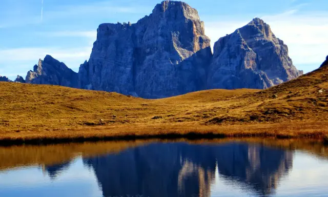 Riflessi maestosi delle Dolomiti nel lago delle Baste, con un prato dorato che separa le acque dalle vette imponenti.