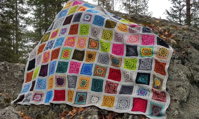 Ett färgglatt, virkat lapptäcke med många olika mönster och färger ligger utspritt över en mossig sten i skogen.