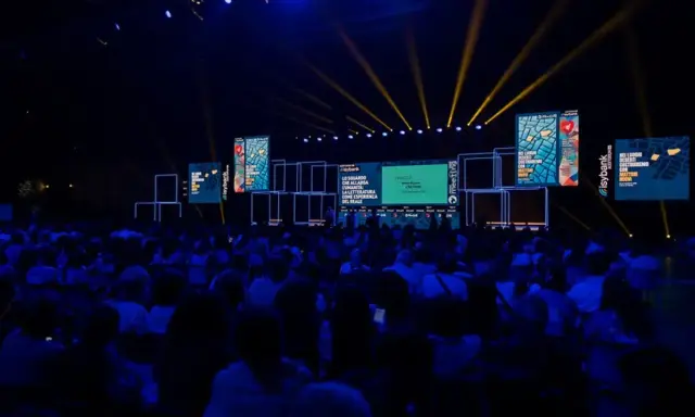 Un grande pubblico partecipa a un meeting per l'amicizia fra i popoli, illuminato da fasci di luce gialla su un palco con schermi.